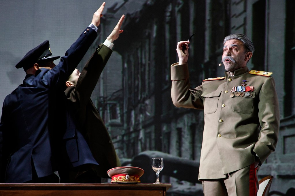 A theatrical performance depicting a historical scene with three male actors. One is dressed in a military uniform with medals, gesturing while speaking. The other two, one in a police uniform and another in civilian attire, are raising their arms in a salute. A table with a military cap and a glass of water is in the foreground.