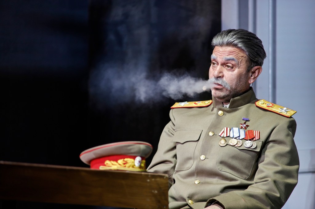 A man in a military uniform with medals sits at a table, exhaling smoke, with a serious expression and a military cap beside him.