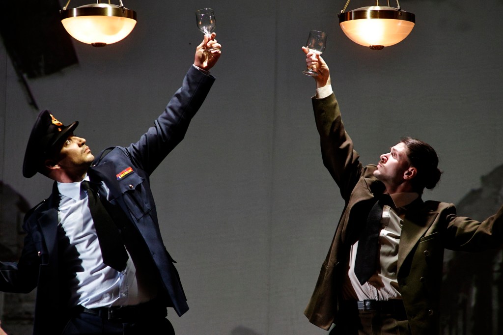 Two actors in a theatrical performance, one dressed as a police officer and the other in a formal suit, both raising glasses in a toast under warm, pendant lighting.
