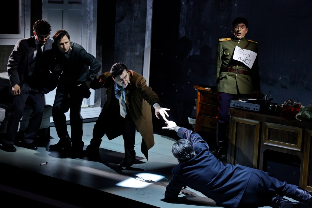 A tense theatrical scene depicting four men in suits engaging in a dramatic struggle, with one reaching out for help while another in military attire holds a sign. The stage is dimly lit, enhancing the dramatic atmosphere.
