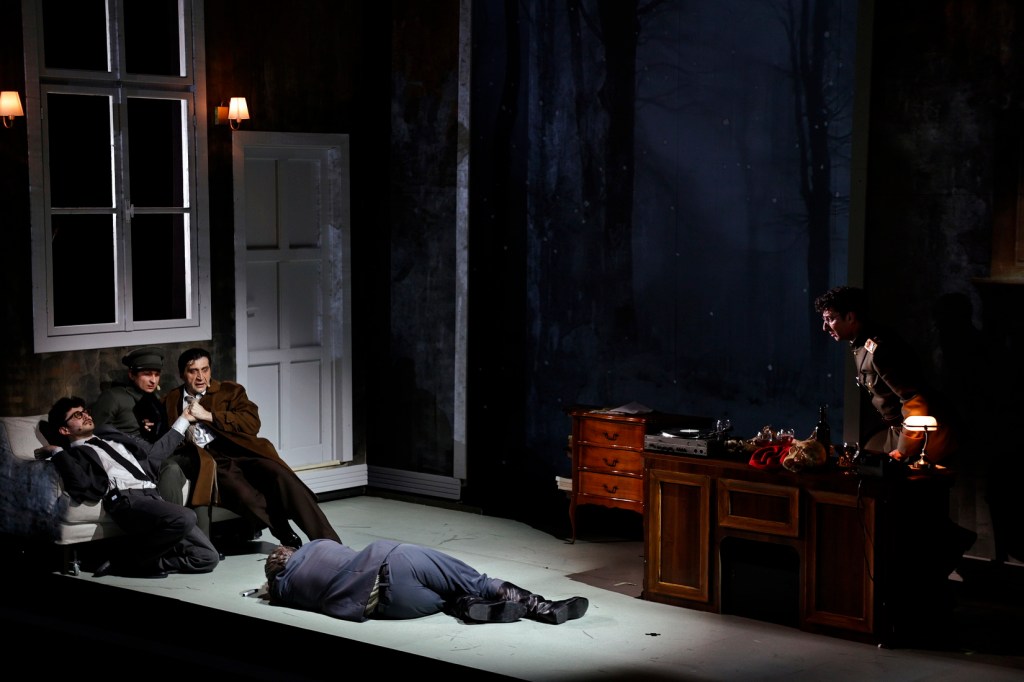 A theatrical scene featuring four characters: three men seated on a couch, appearing distressed, and one man standing near a desk with a record player. A body lies on the floor, adding to the dramatic tension of the setting.