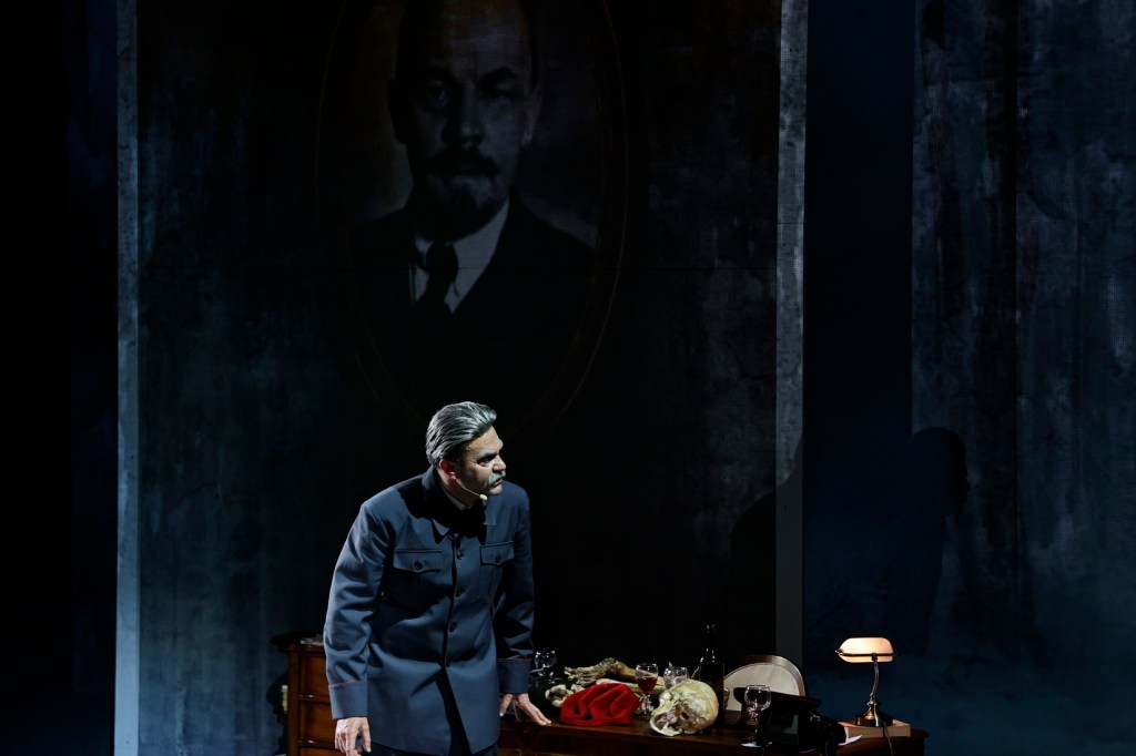 A male actor in a military uniform performs on stage, with a shadowy portrait of a historical figure behind him. The scene includes a wooden table with various props, such as a skull, glasses, and a lamp.