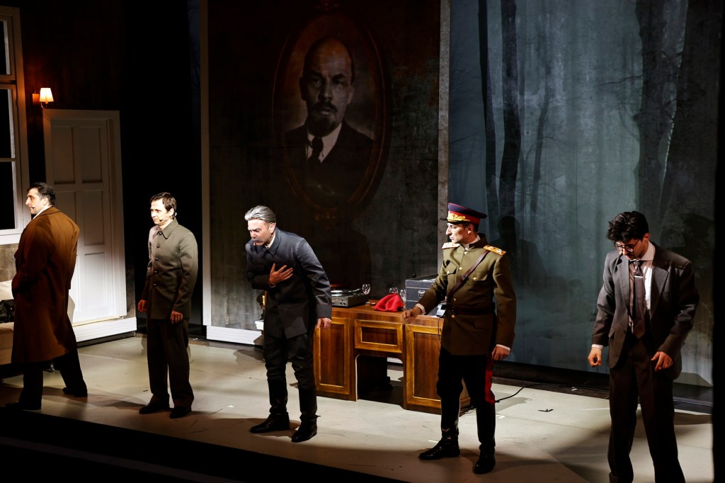 A scene from a theatrical performance featuring five male actors in period costumes, with a backdrop of a portrait of Lenin and dark forest imagery.