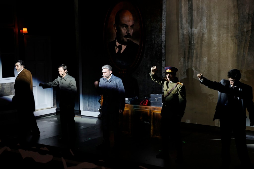 Five actors standing in shadow on stage, with a portrait of Lenin in the background, conveying a dramatic scene.