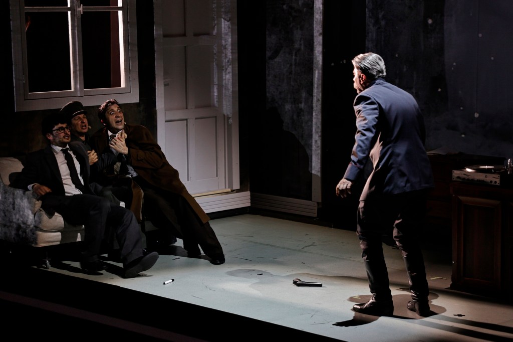 A dramatic scene from a theater performance featuring four male actors. Three actors sit on a couch, displaying expressions of fear and shock, while another actor stands facing them, appearing intense. The setting includes a window and a piece of furniture in the background.