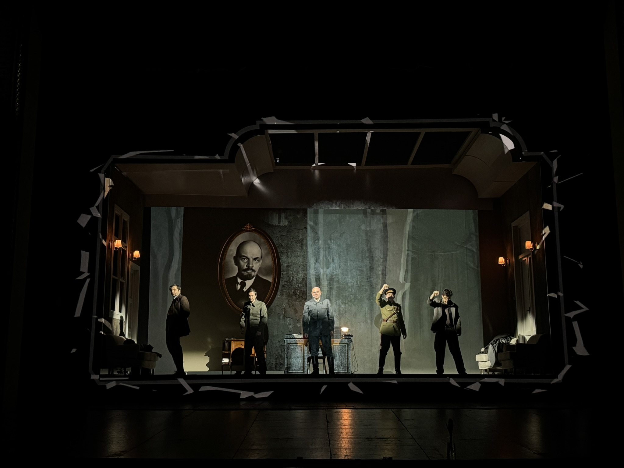 A theatrical scene featuring five actors on stage, with a large portrait of a historical figure in the background. The setting includes furniture and lighting that create an atmospheric interior.