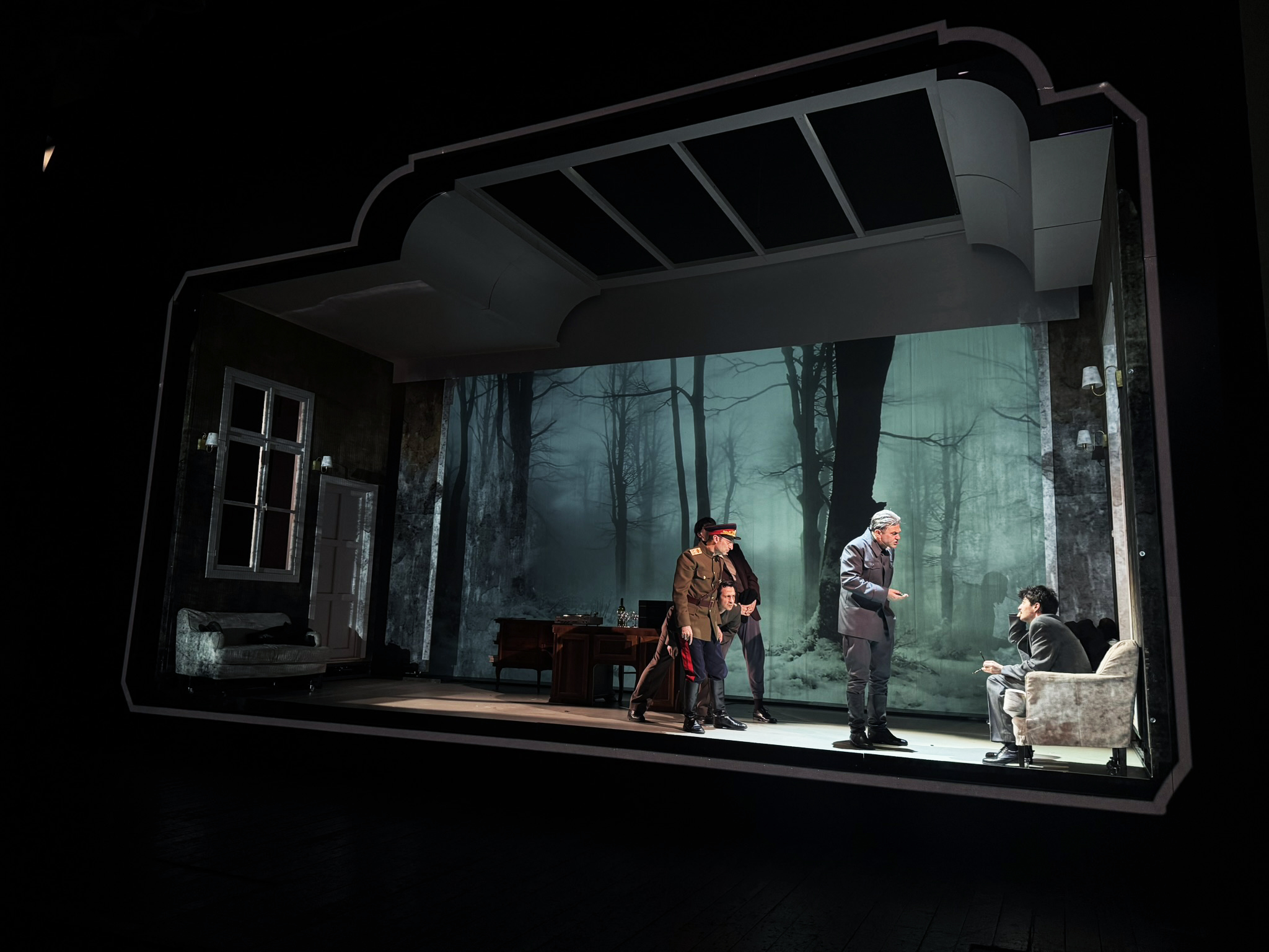A theatrical scene with four actors performing on stage, set against a backdrop of dark, eerie forest imagery. One actor in military attire, another in a suit, engages in conversation with two seated individuals on a couch. The stage features a dimly lit room with furniture, depicting a tense atmosphere.