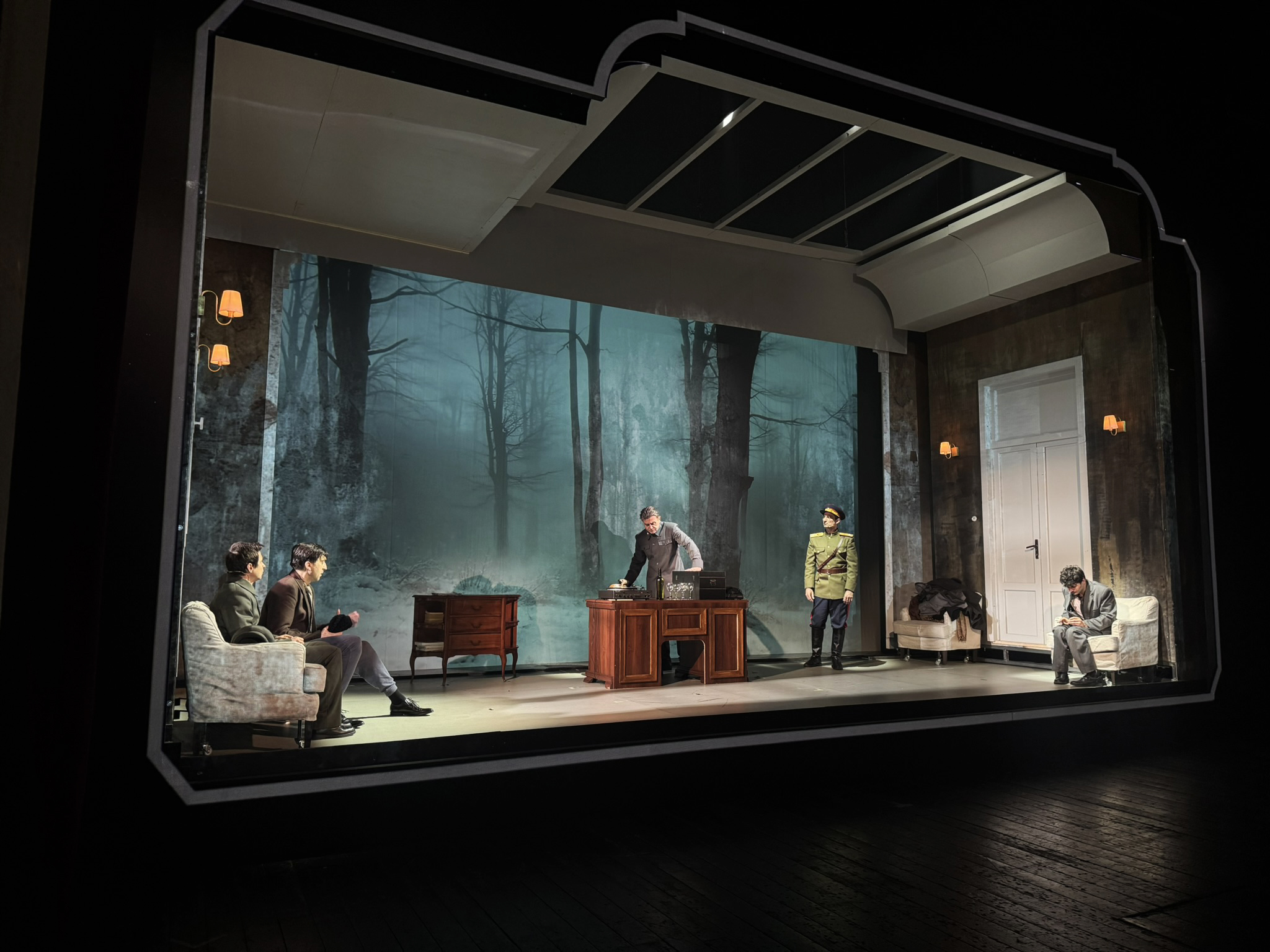 A theatrical scene featuring five characters on stage, set against a backdrop of a foggy forest. Two men are seated on a sofa on the left, while a man in a suit stands at a wooden desk in the center. A soldier in uniform is positioned to the right, and another man sits on a sofa, looking at his phone. The stage is dimly lit with wall sconces illuminating the scene.