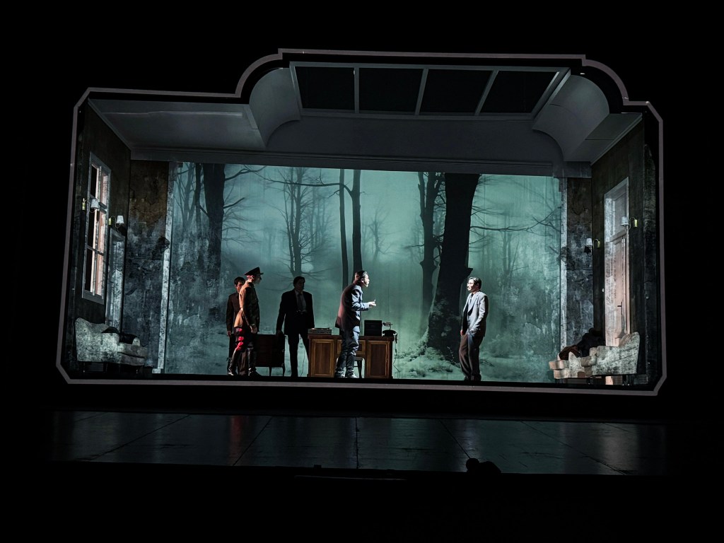 A theatrical scene depicting five actors on stage, interacting in a dimly lit, atmospheric setting with a backdrop of a misty forest visible through windows.