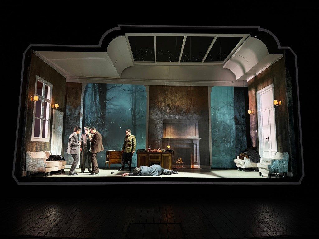 A theatrical scene inside a stylized set featuring five actors engaged in an intense discussion, with a dimly lit room and a backdrop of a forest visible through the windows. One actor lies on the floor, while others stand nearby.