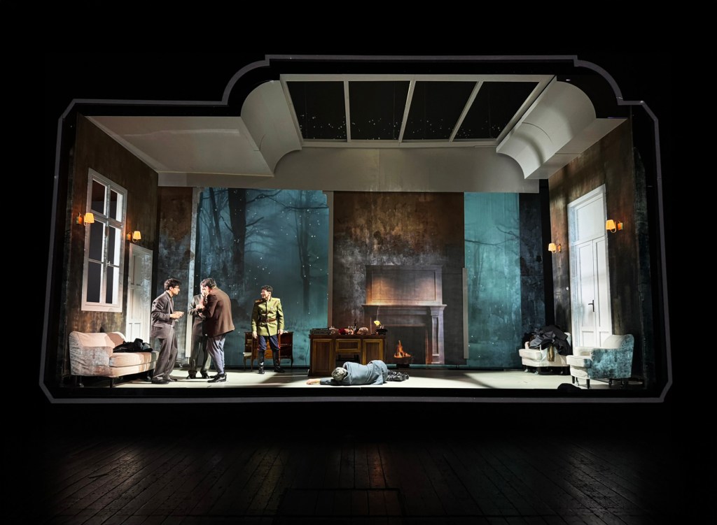 A theatrical scene depicting four characters in a dimly lit room, with a fireplace and an unsettling atmosphere. One character appears to be lying on the floor, with the others engaged in conversation.
