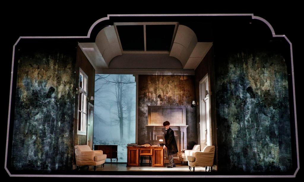 A stage set depicting a room with vintage furniture, including chairs and a table, with a foggy window scene in the background.